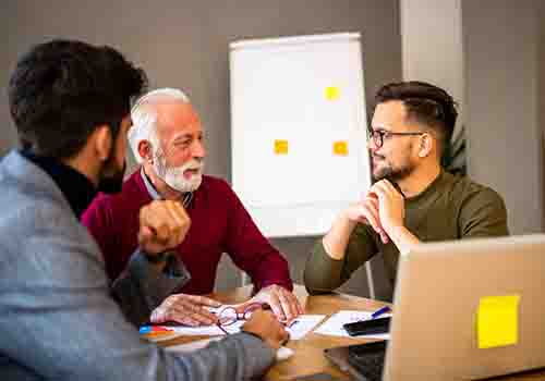 Three men sat around a table with a laptop in the foreground and flipchart in the background