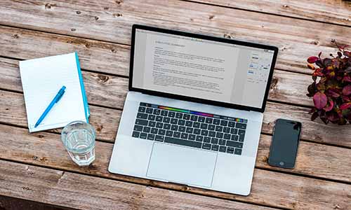 Laptop on a wooden table with glass, notepad and plant to the side