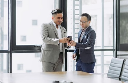 Two men stood in a boardroom talking and sharing something on a phone