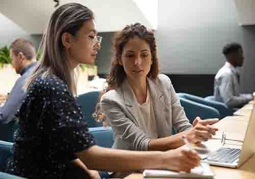 Two women working together at a computer, other people working in the background