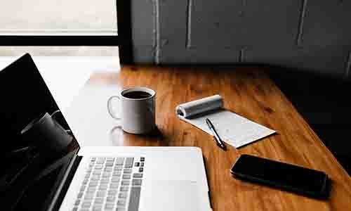 Laptop set up on a desk with a notepad and coffee cup next to it on the table. A window in the background