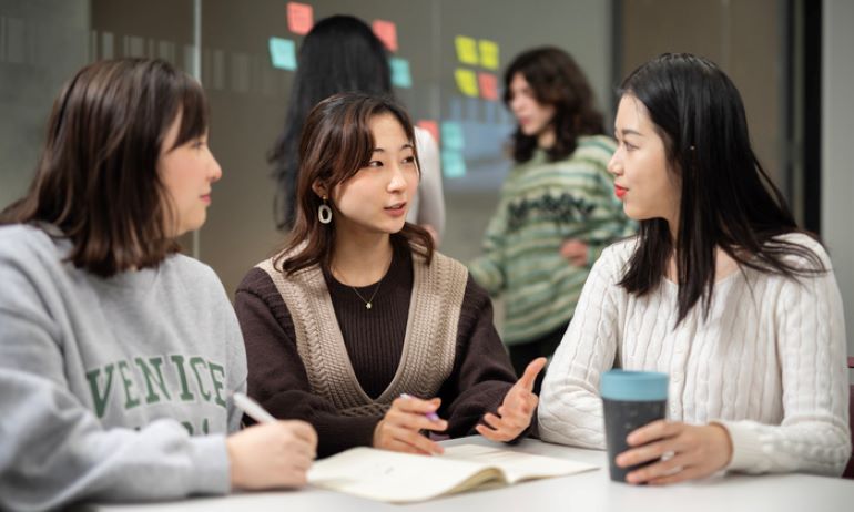 3 Students talking in front of a glass wall dotted with blue, yellow and pink post-it notes.